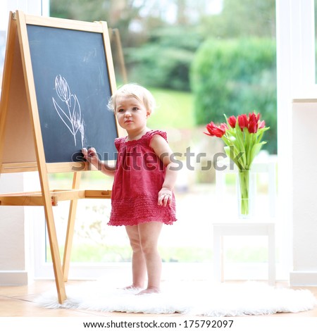 Happy blonde toddler girl in beautiful red dress drawing tulips with chalk on black board standing next to a big window