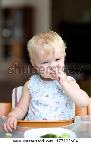 Adorable baby or toddler girl sitting in a high feeding chair biting on delicious freshly cooked broccoli