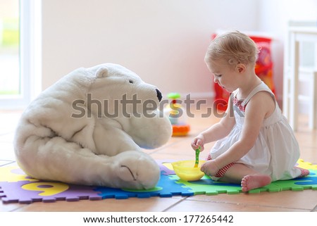 Cute blonde toddler girl feeding her white teddy bear with porridge from spoon sitting indoors at home or kindergarten in a bright room with big window