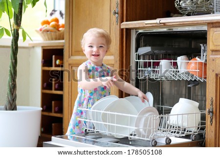 Adorable smiling blonde toddler girl helping in the kitchen taking plates out of dish washing machine