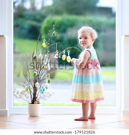 Lovely blonde little toddler girl decorating cherry tree branches with Easter eggs standing indoors next to a big window with street view