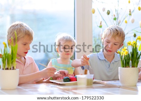 Group of adorable children from one family, two twin brothers and their little toddler sister, decorating and painting Easter eggs sitting together in the kitchen on a sunny day.