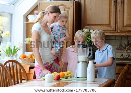 Young mother with three kids, teenager twin sons and little toddler daughter, standing together on sunny kitchen preparing healthy drink with milk and fruits.