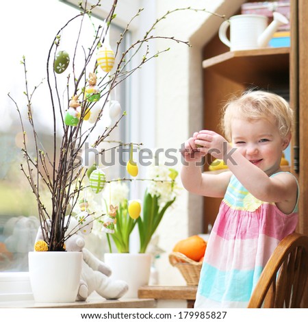 Adorable little blonde toddler girl decorating with Easter eggs cherry tree branches standing in the kitchen next a window with garden view
