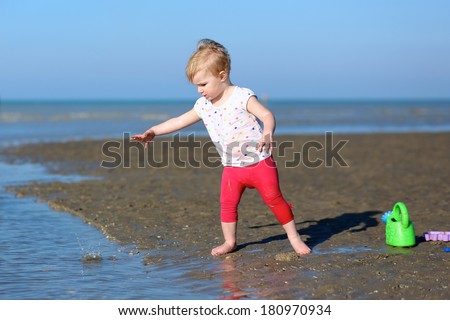 Adorable blonde little toddler girl in colorful outfit plays with watering can at a shore of the sea on a long calm peaceful beach on a warm sunny summer day