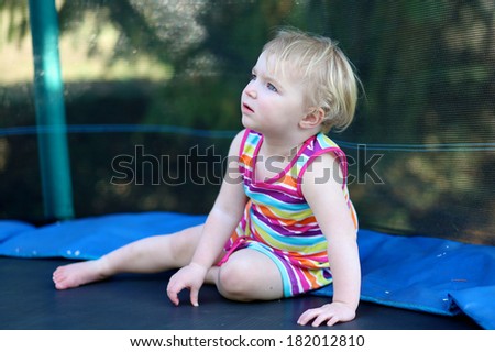 Happy blonde toddler girl in colorful rainbow dress relaxing on trampoline in the garden at the backyard of the house on a sunny summer day