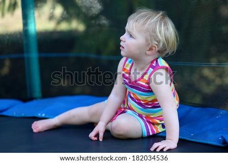 Happy blonde toddler girl in colorful rainbow dress relaxing on trampoline in the garden at the backyard of the house on a sunny summer day