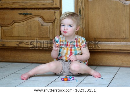 Adorable blonde toddler girl sitting indoors on tiles floor against wooden kitchen cabinetry eating delicious Easter chocolate eggs