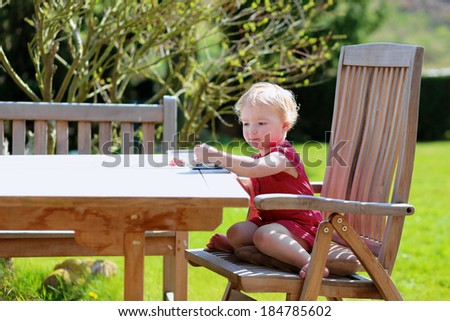 Happy little child, cute blonde toddler girl in beautiful dress snacking outdoors in the garden at the backyard of the house sitting on a wooden teak chair on a sunny summer day eating berries