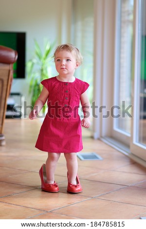 Lovely child, cute funny little toddler girl in beautiful dress trying mom\'s red high heel shoes walking at home along the living room with tiles floor and big windows