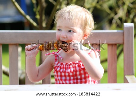Funny child, adorable blonde toddler girl in red dress messy around mouth eating delicious meat made on bbq sitting outdoors in the garden on a wooden chair on a sunny summer day