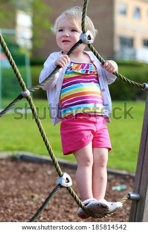 Happy little child, blonde toddler girl in colorful casual suit, having fun on the playground climbing on the net on a sunny summer day