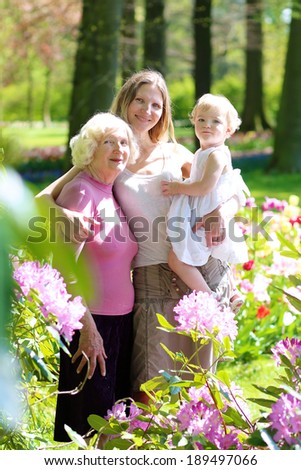 A happy family of three generations, mother, daughter, grandmother and little toddler granddaughter, are standing together in beautiful floral park on a sunny day