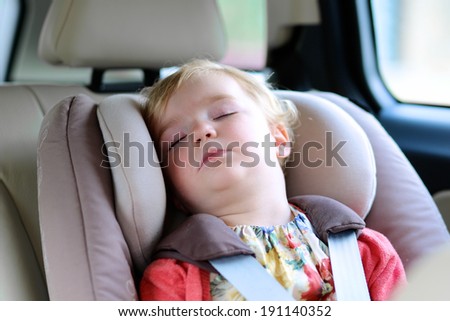 Lovely little child, cute blonde curly toddler girl peacefully sleeping in the car sitting in a baby seat with belt enjoying a family vacation trip in a modern safe vehicle