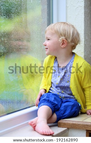 Cute little child, blonde curly toddler girl in colorful casual outfit sitting indoors on a rainy day looking through window with garden view