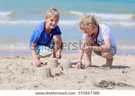 Two happy children, twin brothers, blonde school boys playing on the beach building sand castles on a sunny summer day