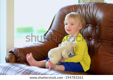 Happy little child, adorable toddler girl with blonde curly hair playing with her teddy bear sitting indoors on brown leather sofa in bright room with big garden view window