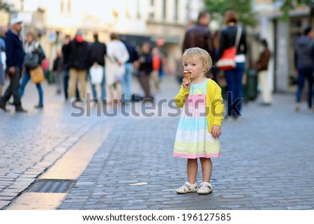 Happy little child, adorable blonde toddler girl in beautiful dress, walking on crowded touristic street in the center of the city eating ice-cream on a sunny summer evening, Brussels, Belgium