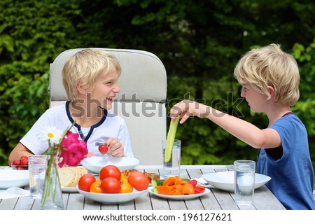 Two happy schoolboys, twin brother, laughing and having healthy lunch or dinner outside sitting at picnic table in the garden at the backyard of the house on a sunny summer day