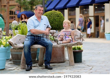 Happy active man relaxing together with his little daughter, adorable blonde toddler girl, sitting on wooden bench in middle of outlet shopping street during summer sales - father and child concept