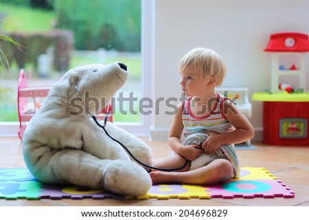Happy child, adorable blonde toddler girl, playing doctor game with her teddy bear sitting comfortable on the floor in playroom at home, school or kindergarten