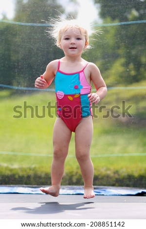 Happy blonde toddler girl in colorful swimming suit jumping on trampoline in the garden at the backyard of the house on a sunny summer day