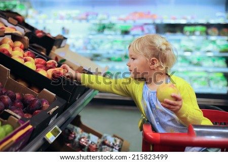 Funny little child, blonde toddler girl, picking fruits from the shelves sitting in the trolley during family shopping in hypermarket