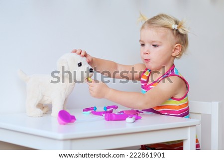 Little child, adorable blonde toddler girl, playing doctor role game with her puppy sitting at small white table in playroom at home, school or kindergarten