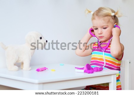 Adorable blonde toddler girl playing doctor role game putting on stethoscope in order to examinate her puppy sitting at small white table in playroom at home, school or kindergarten