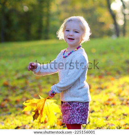 Happy little child, cute toddler girl with curly blonde hair wearing yellow jacket playing with colorful leaves in park on sunny warm autumn day