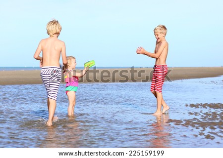 Group of three happy active kids, 2 twin brothers and little sister, cute toddler girl in colorful swimming suit, playing on the beach, running and splashing with the water guns at low tide