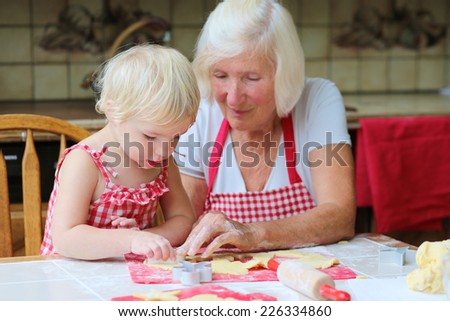 Loving caring grandmother, beautiful senior woman, baking tasty sweet cookies together with her granddaughter, cute little toddler girl, sitting at the table in classic traditional wooden kitchen