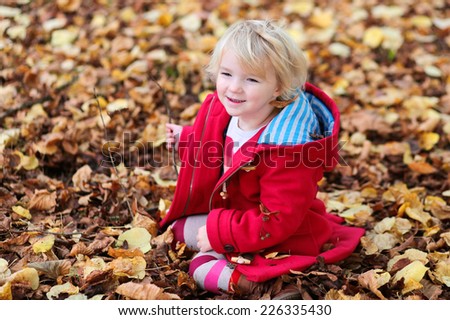 Adorable little child, blonde toddler girl in warm red duffle coat, playing with leaves in autumn park