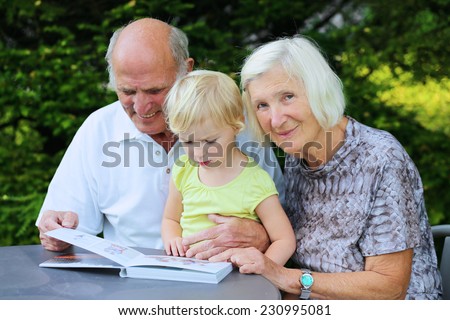 Happy smiling grandparents relaxing outdoors in the garden watching with their granddaughter photo book, a gift from children as a memory of summer vacation