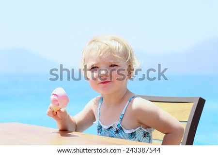 Happy healthy little child, cute blonde toddler girl, enjoying summer vacations eating delicious strawberry ice cream sitting at beach restaurant with sea view in tropical resort - summertime concept