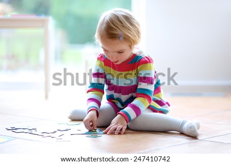 Adorable little child, blonde toddler girl, playing with puzzles sitting on the tiles floor next to a big window at home or kindergarten