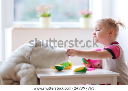 Adorable smiling child, blonde toddler girl, playing at home or kindergarten, cutting plastic vegetables with knife and feeding her toys, a doll and a teddy bear. Promoting healthy lifestyle for kids.