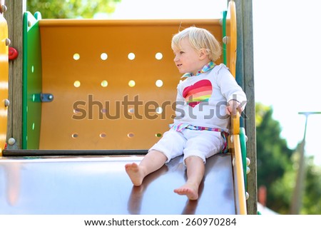 Funny little child, adorable preschooler girl in pretty dress having fun on a slide in the park on summer day