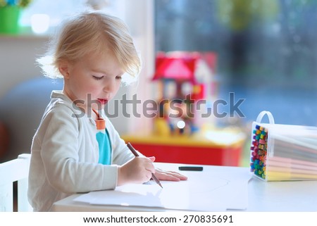 Cute little child, blonde preschooler girl is drawing and painting with colorful felt-tip pens at home or kindergarten sitting at small table in bright sunny playroom