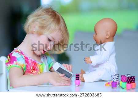 Little child, cute toddler girl having fun playing at home with colorful nail polish doing manicure and painting nails to her doll