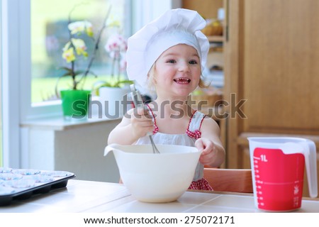 Baking with children: little happy kid, adorable toddler girl in white chef hat mixing dough ingredients in bowl helping mother to prepare delicious pastry in the kitchen