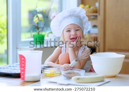 Baking with children: little happy kid, adorable toddler girl in white chef hat helping mother to prepare delicious muffins in the kitchen. Bowl, flour, butter and banana on the table.