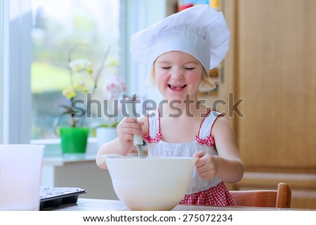 Baking with children: little happy kid, adorable toddler girl in white chef hat mixing dough ingredients in bowl helping mother to prepare delicious pastry in the kitchen