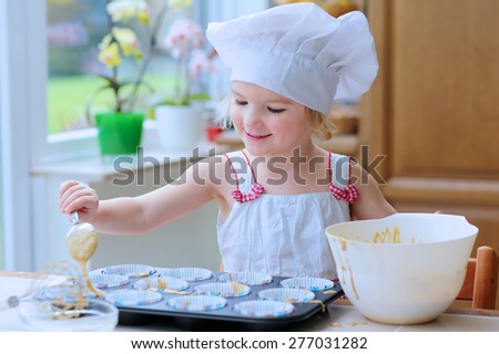 Baking with children. Little happy kid, adorable toddler girl in white chef hat filling cupcakes form with dough ingredients helping mother to prepare delicious pastry in the kitchen