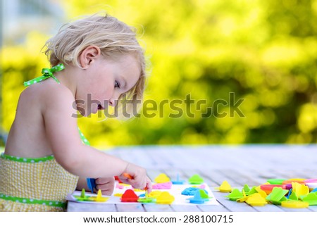 Little girl playing with plasticine and colorful forms. Happy child, adorable toddler girl creating from modeling compound dough, sitting outdoors in the garden on sunny summer day