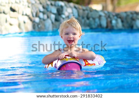 Adorable happy little child, curly toddler girl in swimming suit having fun relaxing and floating on an inflatable toy ring in a pool on sunny day during summer vacation in resort