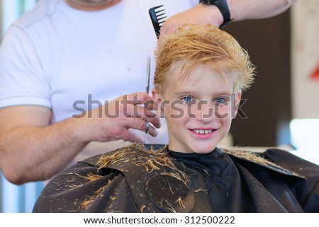 Teenage boy having a haircut in a salon. Cute caucasian teenager sitting at the hairdresser. Barber holding in his hand hairbrush and scissors.