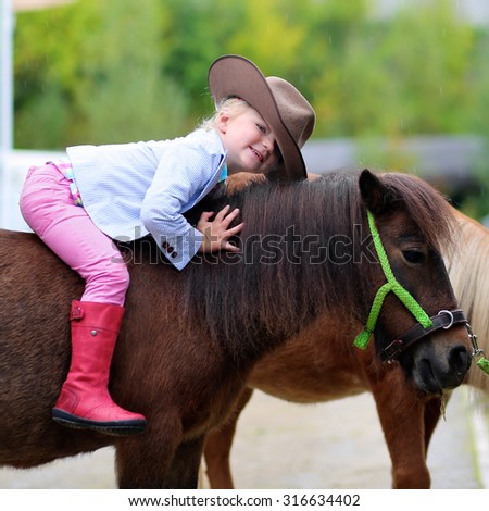 Lovely cowgirl sits on little pony horse in the farm. Pretty preschooler girl wearing cowboy hat playing with animals outdoors on sunny day.