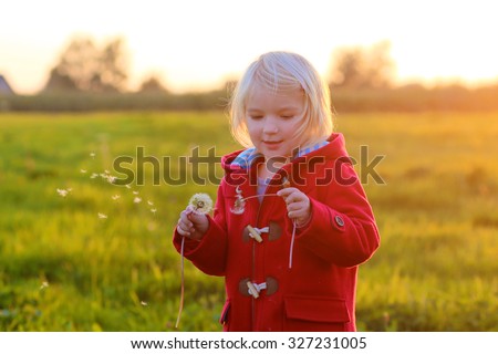 Little girl wearing red coat playing with dandelions outdoors in the countryside at sunset. Cute child enjoying nature on beautiful autumn or spring evening.