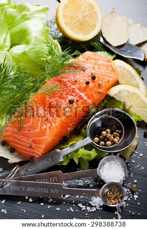 Delicious salmon fillet, rich in omega 3 oil, aromatic spices in measuring spoons and lemon on fresh lettuce leaves on black wooden background. Healthy food, diet and cooking background.